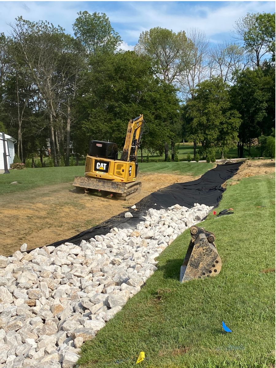 Mini excavator trenching a rock drain line across a yard